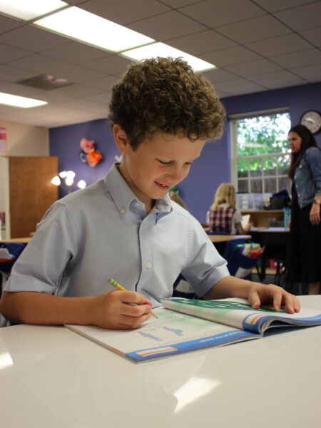 A young boy sitting at a table with a book in a classroom at River Oaks Baptist School