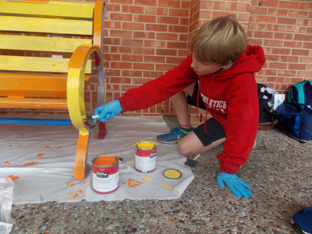 A young boy painting a bench in front of a brick wall at River Oaks Baptist School