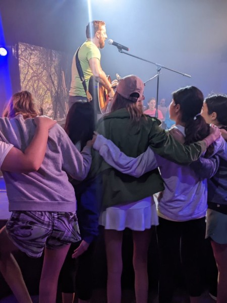 A group of people standing around a man on a music concert stage at River Oaks Baptist School