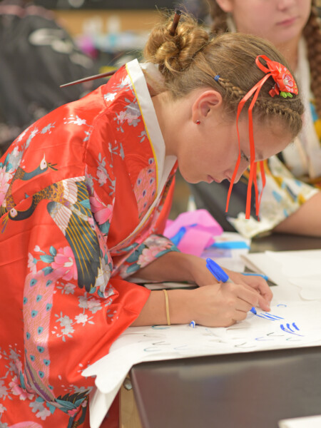 A student in a red kimono writing on a piece of paper in a classroom at River Oaks Baptist School