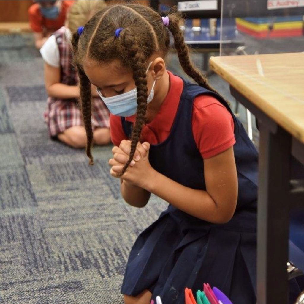 A little girl sitting on the floor wearing a face mask and praying at River Oaks Baptist School