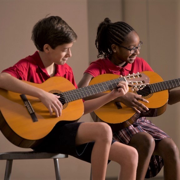 A couple of kids sitting next to each other playing guitars at River Oaks Baptist School